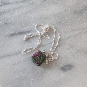 May include: A silver necklace with a square gemstone pendant. The gemstone displays a mix of green and pink colours. The chain has a delicate, twisted design, and the clasp is visible.