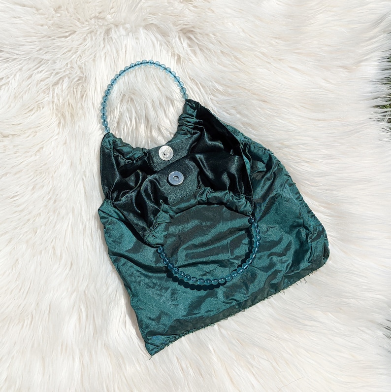 Pode incluir: Uma bolsa de m&atilde;o de cetim azul-esverdeado com uma al&ccedil;a de contas. A bolsa tem um design franzido e um fecho de aba.