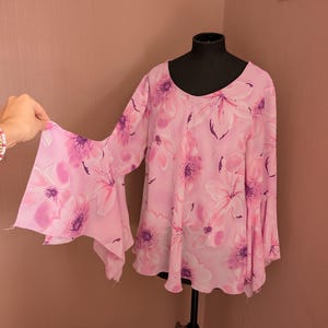 May include: A pink floral blouse with bell sleeves is displayed on a black mannequin. The blouse has a scoop neck and a pattern of large, light pink flowers with purple centres and accents. The fabric appears to be lightweight and flowing.