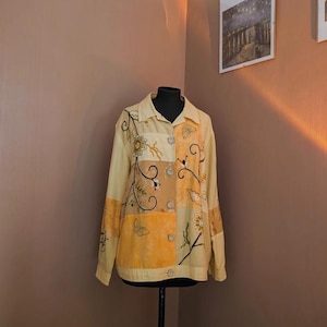 Puede incluir: Una camisa amarilla y naranja bordada con un patrón floral. La camisa tiene cuello y mangas largas.