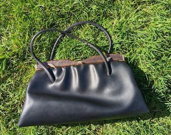Puede incluir: Un bolso de mano de cuero negro con un marco de madera marrón y un cierre de metal. El bolso está sobre un lecho de hierba verde.