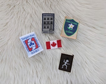 Könnte beinhalten: Fünf gestickte Aufnäher, darunter eine kanadische Flagge, ein Girl Guides-Aufnäher mit einer weißen Blume und dem Text "Girl Guides Quebec", ein Aufnäher mit dem Text "Sangam Area Guides Canada Quebec" und einem Kleeblatt, ein Aufnäher mit dem Text "Guide House Montreal, Quebec" und einem Gebäude sowie ein Aufnäher mit einer Strichmännchenfigur, die eine Axt hält.