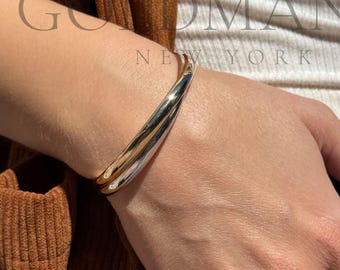 May include: A gold and silver-toned bracelet with a curved design. A gold ring is on the finger. The image also shows a person wearing a white top, blue jeans, and a brown corduroy jacket. The text "GOMANIA NEW YORK" is visible.