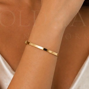 May include: A gold-colored, rectangular bangle bracelet. The bracelet is worn on the arm and has a simple, elegant design. The clasp is visible. The background is a woman's skin and a white satin top.
