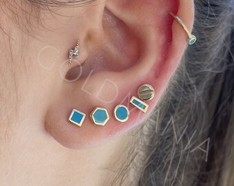 May include: A close-up of a person's ear with multiple small earrings. The earrings are gold with blue enamel and are in the shapes of a square, hexagon, circle, rectangle, and a screw.