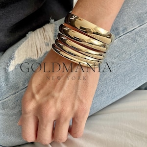 May include: A set of five gold-toned bangle bracelets stacked on a person's wrist. The bracelets are all smooth and have a shiny finish. The bracelets are made of metal and are a popular fashion accessory.