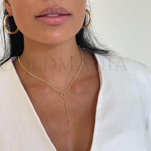 Könnte beinhalten: Eine goldfarbene Halskette mit einem Y-förmigen Anhänger und passenden Creolen. Die Halskette hat ein filigranes Kettendesign mit einem kleinen kreisförmigen Anhänger. Die Ohrringe sind groß und kreisförmig. Das Model trägt ein weißes Oberteil.