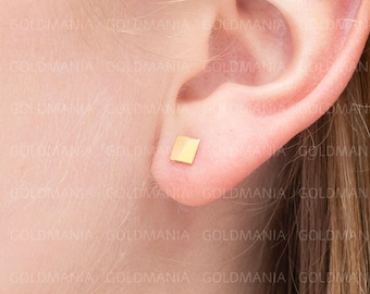 May include: A close-up of a gold square stud earring on an ear. The earring is a small, geometric shape, and the gold color is bright and reflective. The ear is in soft focus, with the earring as the focal point.