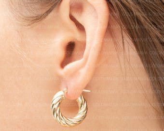 May include: A close-up of a gold hoop earring with a twisted rope design. The earring is worn on an ear, with the focus on the jewelry's texture and shine. The earring is a classic accessory.