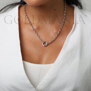 May include: A silver-colored chain necklace with a circular pendant. The necklace is worn against a white shirt. The brand name "GOLDMANIA NEW YORK" is visible in the background.