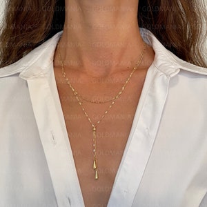 Könnte beinhalten: Eine goldene mehrreihige Halskette mit einer zarten Kette und einem Anhänger mit zwei kleinen, spitzen Tropfen. Die Kette wird von einer Person mit einem weißen Hemd getragen.