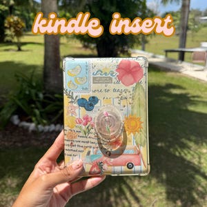 May include: A clear Kindle insert with a collage design featuring lemons, flowers, and text. The insert has a pop-up grip in the center. The words "kindle insert" are written in a cursive font across the top.