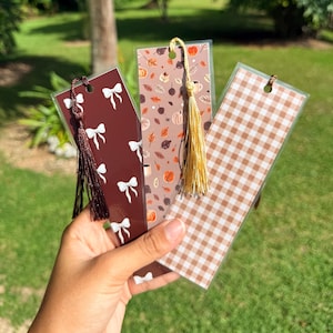 May include: Three decorative bookmarks with tassels. One is dark brown with white bows, another is tan with autumn leaves and pumpkins, and the third is tan and white gingham. The bookmarks are held in front of a green background.