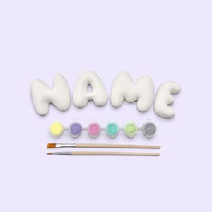 May include: A white ceramic word sculpture spelling the word NAME in a rounded, bubble-like font. Below the sculpture is a plastic strip containing six small circular paint pots in yellow, purple, pink, teal, light green, and gray, plus two brushes.