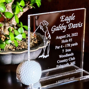 May include: A clear acrylic plaque with an engraved image of a golfer and the text "Eagle Gabby Davis August 24, 2022 Hole #5 Par 4 - 178 yards 7 Iron Woodlands Country Club Falmouth, ME". A white golf ball is in the foreground.