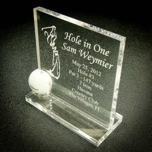 May include: A clear acrylic plaque with a golfer silhouette and a golf ball. The plaque reads "Hole in One Sam Weymier May 25, 2012 Hole #3 Par 3 - 147 yards 7 Iron Havana Country Club The Villages, FL"