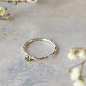 May include: A thin, polished silver band ring featuring a single, small, round, bezel-set green gemstone. The ring has a smooth, circular profile and a bright, reflective metallic finish, showcasing a simple design with a vibrant green accent stone.