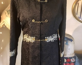 May include: A black brocade jacket with gold and silver embroidery. The jacket has a mandarin collar, long sleeves, and a fitted waist. It features a decorative gold button closure and a silver embroidered trim around the collar and cuffs.