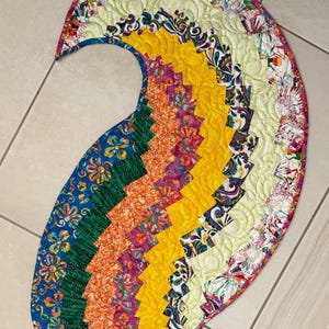 May include: A colorful, quilted paisley-shaped table runner. The runner features a patchwork design with vibrant hues of blue, green, orange, yellow, and purple. The edges are finished with a floral print.