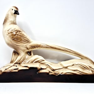 May include: A ceramic pheasant figurine in shades of cream and brown. The bird is perched on a stylized rock formation with a dark brown base. The pheasant has a long, flowing tail and detailed feather patterns.