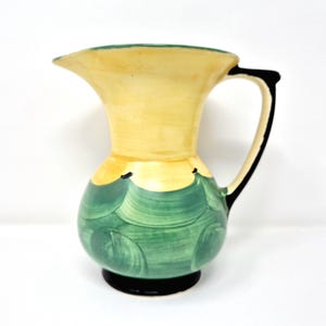 May include: A vintage ceramic pitcher with a yellow top and a green, wave-like pattern on the body. The handle is black, and the base is also black. The pitcher has a spout for easy pouring and is likely used for serving beverages.