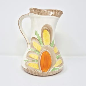 May include: A vintage ceramic pitcher with a white base and a brown handle. The pitcher is decorated with a stylized floral design in shades of yellow, orange, and brown. The rim of the pitcher is also brown.