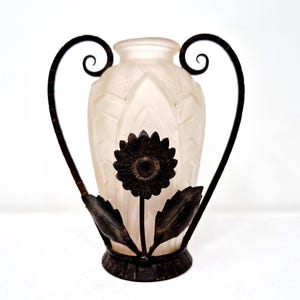 May include: A frosted glass vase with a dark metal floral design. The vase has a sunflower in the center and is framed by curved metal handles. The vase is approximately 10 inches tall.