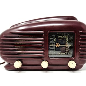 May include: A dark maroon, streamlined radio with a horizontal ribbed design and three cream-coloured circular control knobs along the bottom. The front features a rectangular glass dial with printed city names and a central gold-coloured indicator needle.