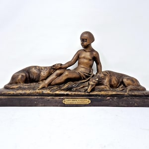May include: A bronze sculpture depicting a young person seated with two dogs. The sculpture has a dark brown base and a plaque with text. The figures are detailed and the overall style is classical.