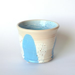 May include: A small, white ceramic planter with blue and white paint splatters. The planter has a rounded bottom and a flared top.