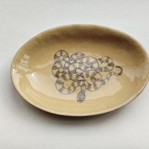 May include: A light brown oval ceramic dish with a black and white snake design. The snake is coiled and has a striped pattern.