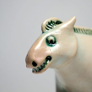 Pode incluir: Uma estatueta de cavalo em cerâmica com um esmalte brilhante em tom branco sujo e detalhes verdes distintos na crina, olhos e narinas. A escultura apresenta uma cabeça estilizada com boca aberta, dentes visíveis e um acabamento liso e reflexivo.