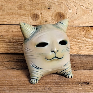 Könnte beinhalten: Eine Keramik-Katzenfigur mit weißer und blauer Glasur. Die Katze hat einen breiten, lächelnden Mund und große, schwarze Augen. Die Figur steht auf einer Holzoberfläche.