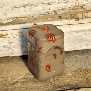 May include: A small, square, lidded box with a textured, mottled gray surface. The box is adorned with orange, leaf-like designs. The box is sitting on a weathered wooden surface.