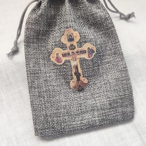 May include: A gray drawstring bag with a decorative wooden cross affixed to the front. The cross features religious iconography and is made of light-colored wood. The bag is made of a textured, woven material.