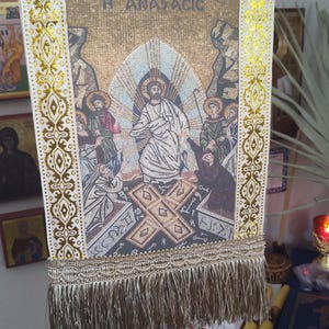 May include: A rectangular religious banner with a mosaic-style image of Jesus Christ surrounded by figures. The banner has a gold and white decorative border with fringe at the bottom. The text "H ANACTACIC" is visible on the banner.