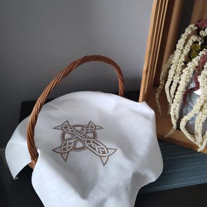 May include: A woven wicker basket with a curved handle, covered by a white cloth. The cloth features a brown Celtic knot design. The basket is near a wooden crate with dried flowers.