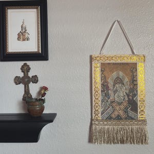 May include: A framed religious tapestry with a gold border and fringe hangs on a wall. A framed church drawing and a decorative cross with a small pot of flowers are also displayed.