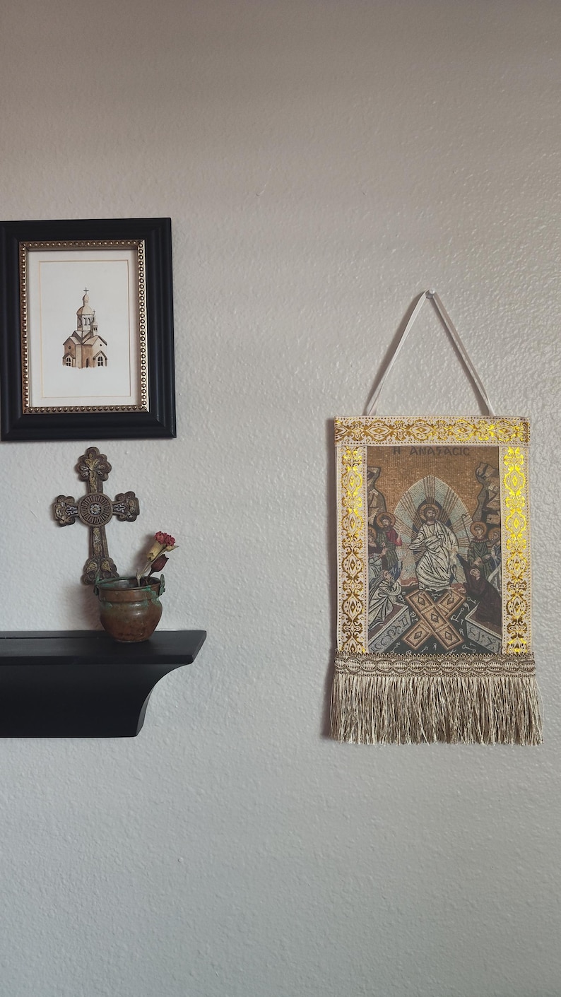 May include: A framed religious tapestry with a gold border and fringe hangs on a wall. A framed church drawing and a decorative cross with a small pot of flowers are also displayed.