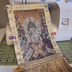 May include: A tapestry with a religious scene, featuring figures and a cross, is held in place by clips on a sewing machine. The tapestry has gold trim, fringe, and the word "ANATACIC". The background is a white sewing machine.