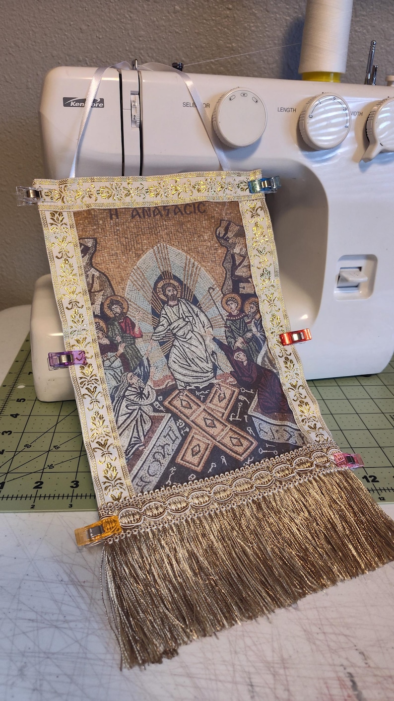 May include: A tapestry with a religious scene, featuring figures and a cross, is held in place by clips on a sewing machine. The tapestry has gold trim, fringe, and the word "ANATACIC". The background is a white sewing machine.