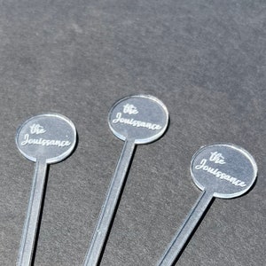 May include: Three clear acrylic cocktail stirrers with a round, silver-colored top. Each top has the text "The Jouissance" engraved on it.