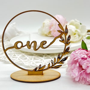 May include: Wooden table number with a circle design and the word "one" in a cursive font. The circle is decorated with a leafy branch design. The table number is on a wooden base.