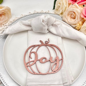 May include: A rose gold pumpkin-shaped place card with the word "Joy" written inside. The place card is on a white napkin on a silver charger.