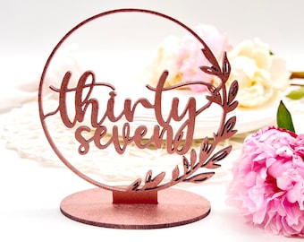 May include: Rose gold cake topper with the words "thirty seven" in a cursive font, surrounded by a circle and leaves.