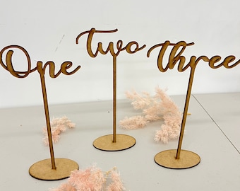May include: Three wooden table number signs with the words "One", "Two", and "Three" written in a cursive font. The signs are on small wooden bases.