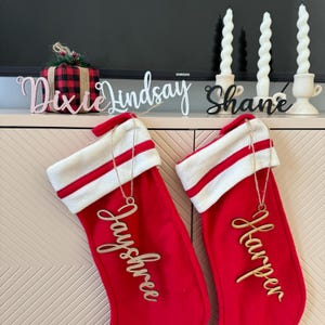 May include: Two red Christmas stockings with white cuffs and red and white stripes hang on a beige cabinet. Each stocking has a wooden name tag hanging from it. The stocking on the left has the name "Jayshree" and the stocking on the right has the name "Harper".