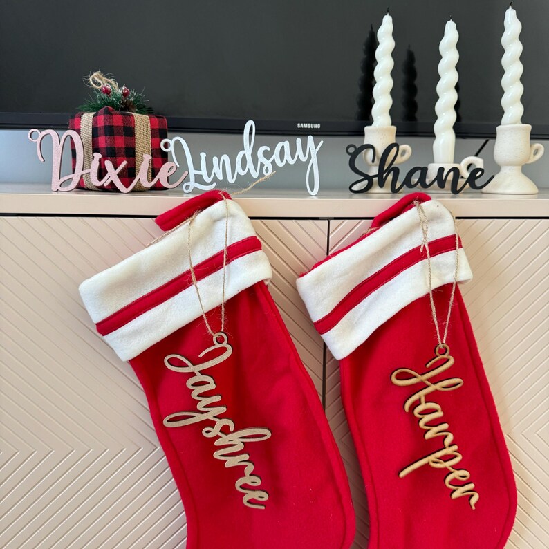 May include: Two red Christmas stockings with white cuffs and red and white stripes hang on a beige cabinet. Each stocking has a wooden name tag hanging from it. The stocking on the left has the name "Jayshree" and the stocking on the right has the name "Harper".