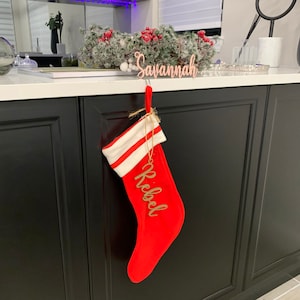 May include: A red Christmas stocking with a white cuff is hanging from a hook. The stocking has a gold tag with the name "Rebbec" written on it. A wooden sign with the name "Savannah" is hanging above the stocking.