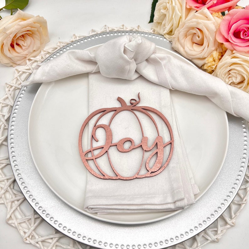 May include: A rose gold pumpkin-shaped place card with the word "Joy" written inside. The place card is on a white napkin on a silver charger.